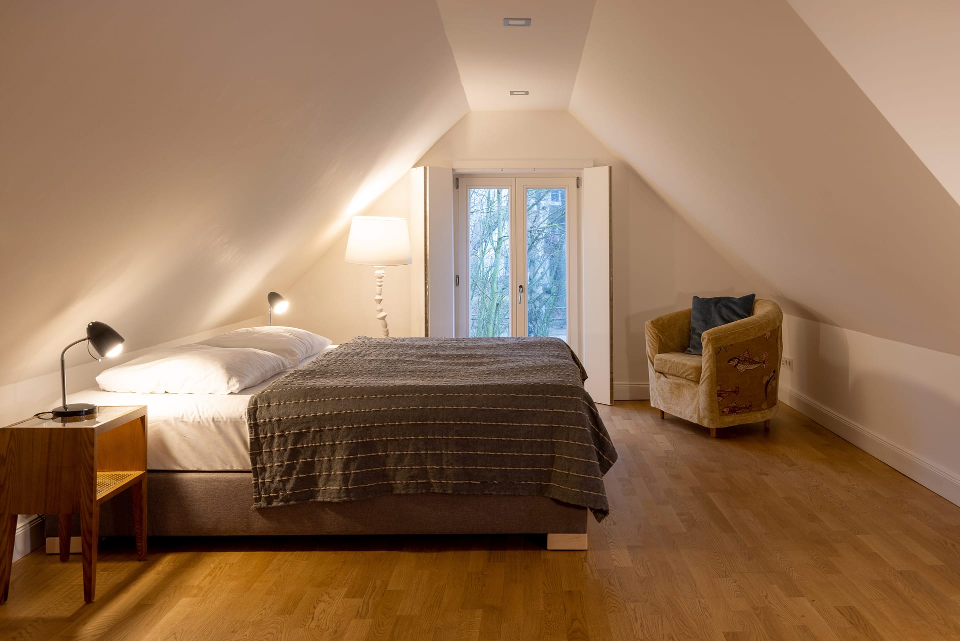 Ein Schlafzimmer unter einer Dachschräge mit grauem Boxspringbett, Holzboden, einem beigen Sessel und direktem Blick auf ein Balkonfenster.