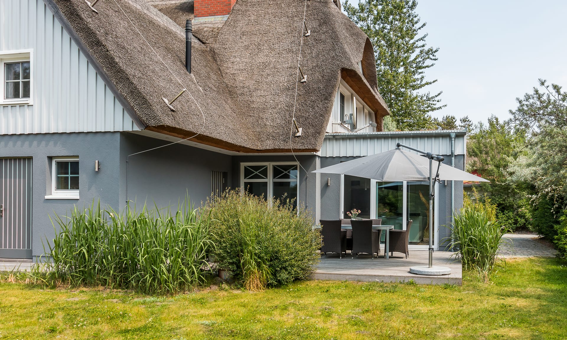 Ein reetgedecktes Haus mit grauer Fassade, Holzterrasse, Sonnenschirm und Gartenmöbeln auf einer Rasenfläche.