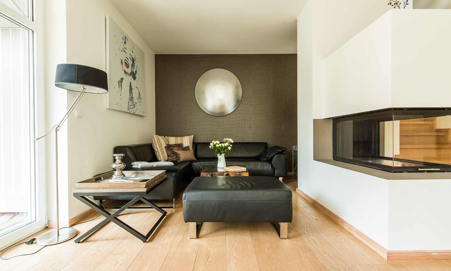 Wohnzimmer mit schwarzer Lederecke, Holzboden, einem Kamin und einer markanten silbernen Wanddekoration.