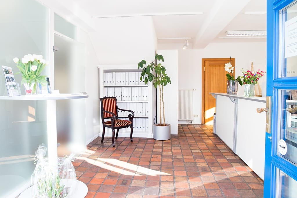 Eingang zum Büro: links ein Tisch mit Blumenvasen und Flyern, geradeaus ein Regal mit Ordnern, rechts eine Theke, auf der Blumen stehen.