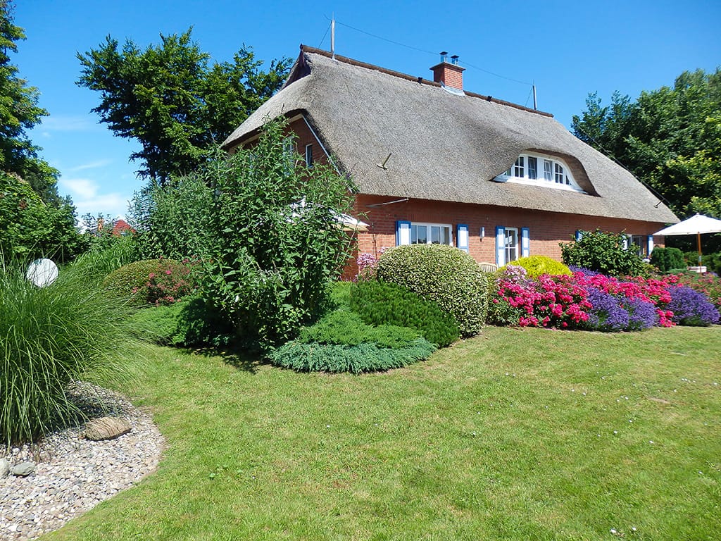 Reetdachhaus im Garten mit bunte Blumen und Sträuchern, sonniges Wetter, gepflegtes Grundstück, blauer Himmel.