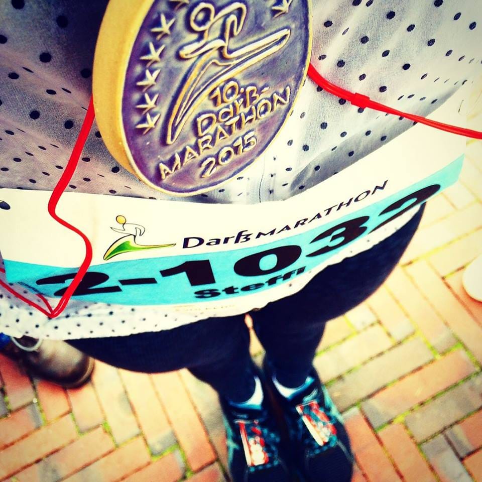 Eine Medaille vom „10. Darß-Marathon 2015“ und eine Startnummer mit dem Namen „Steffi“ an einer Läuferin auf einem gepflasterten Weg.