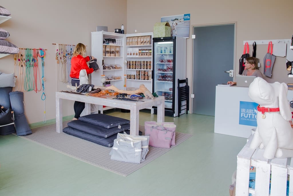 Zwei Frauen in einem hellen Hundegeschäft namens Urlaubsfutter, das Hundezubehör und Futter anbietet, wobei eine am Tresen und die andere an einem Warentisch steht.