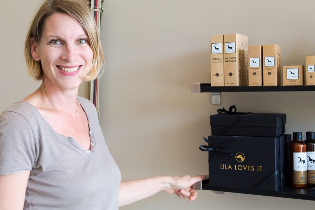 Eine lächelnde Frau steht in einem Hundepflegeshop, umgeben von braunen Kartons und schwarzen Schachteln mit Pflegeprodukten für Hunde.