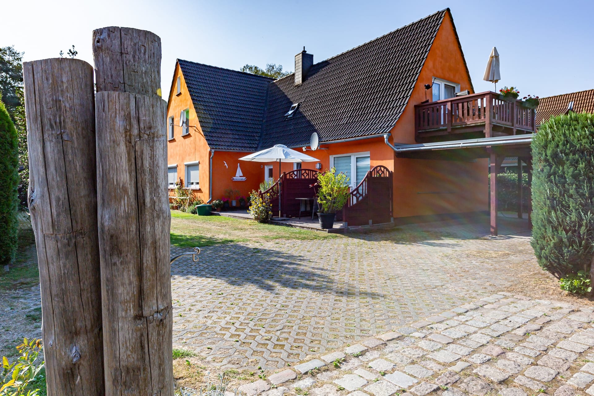 Ein oranges Haus mit dunklem Ziegeldach, einem Holzbalkon, einer Außentreppe und gepflasterten Wegen auf dem Grundstück.