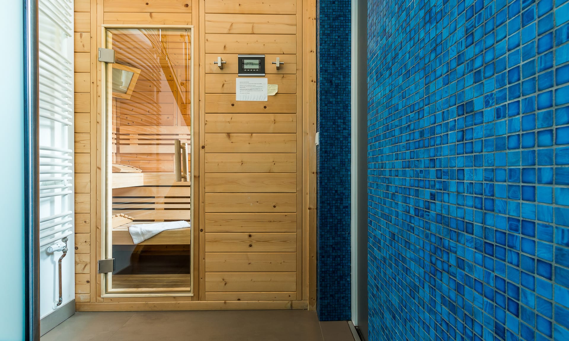 Eine Holzsauna mit Glastür neben einer Wand mit blauen Mosaikfliesen und einem weißen Handtuchwärmer.