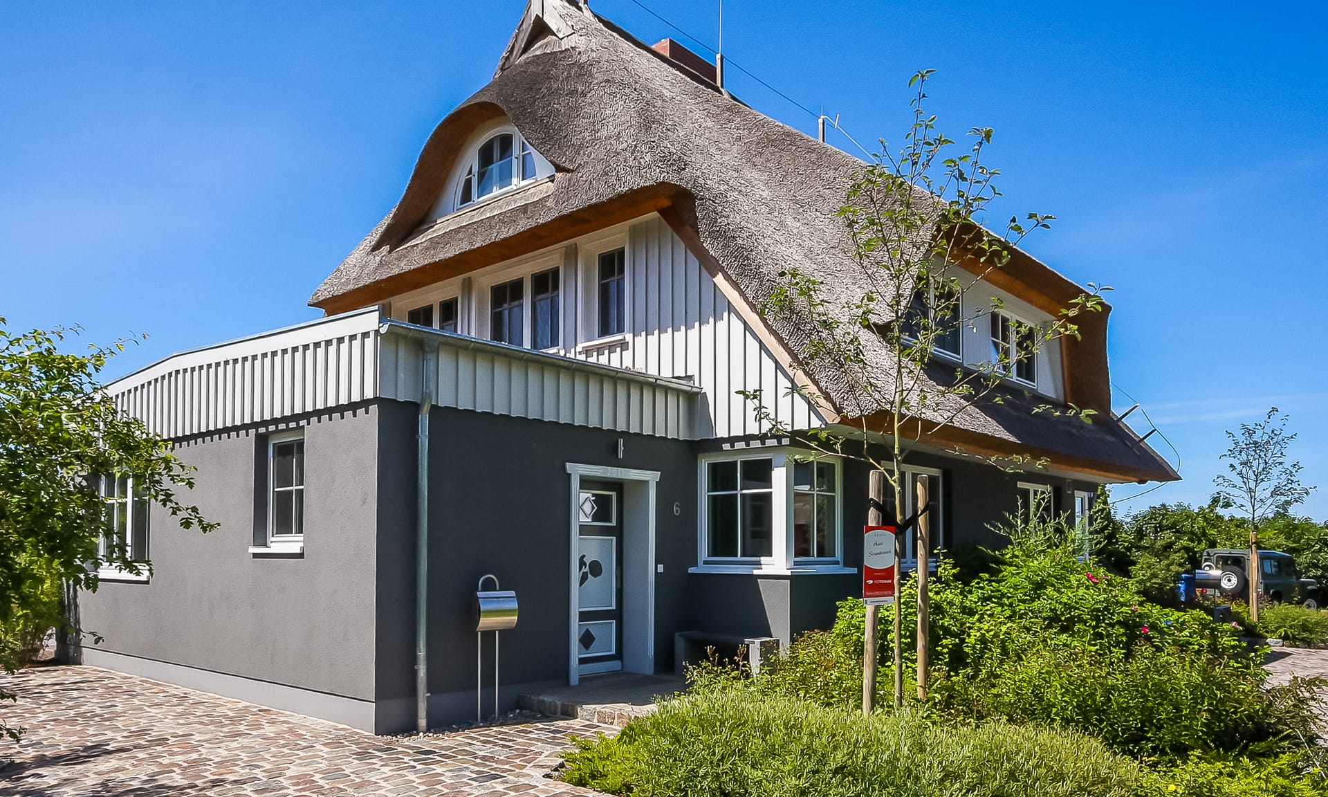 Ein modernes, reetgedecktes Einfamilienhaus mit dunkelgrauem Erdgeschoss und hellgrauem Obergeschoss unter blauem Himmel.