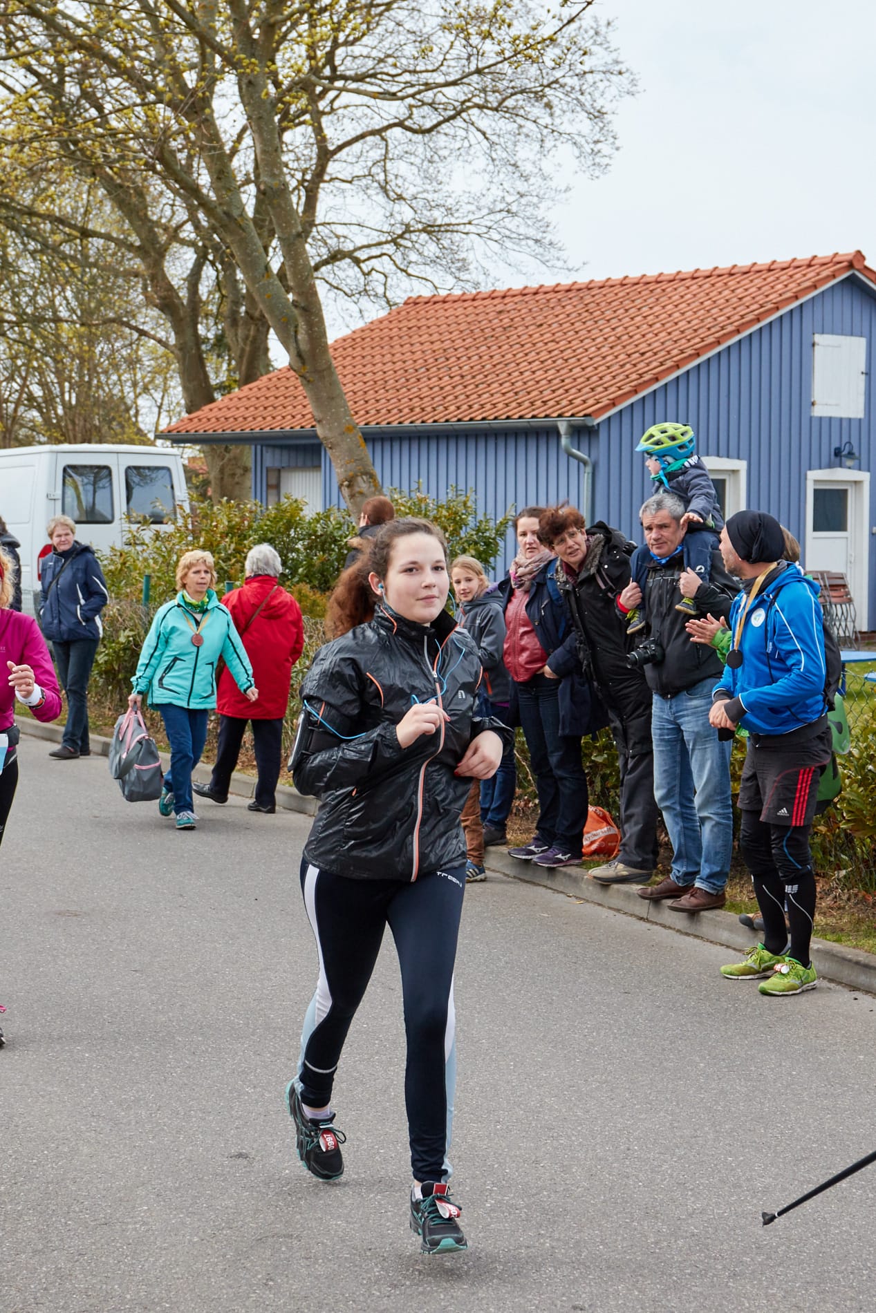 Eine junge Läuferin in schwarzer Sportkleidung auf einer asphaltierten Straße vor Zuschauern und einem blauen Holzhaus.