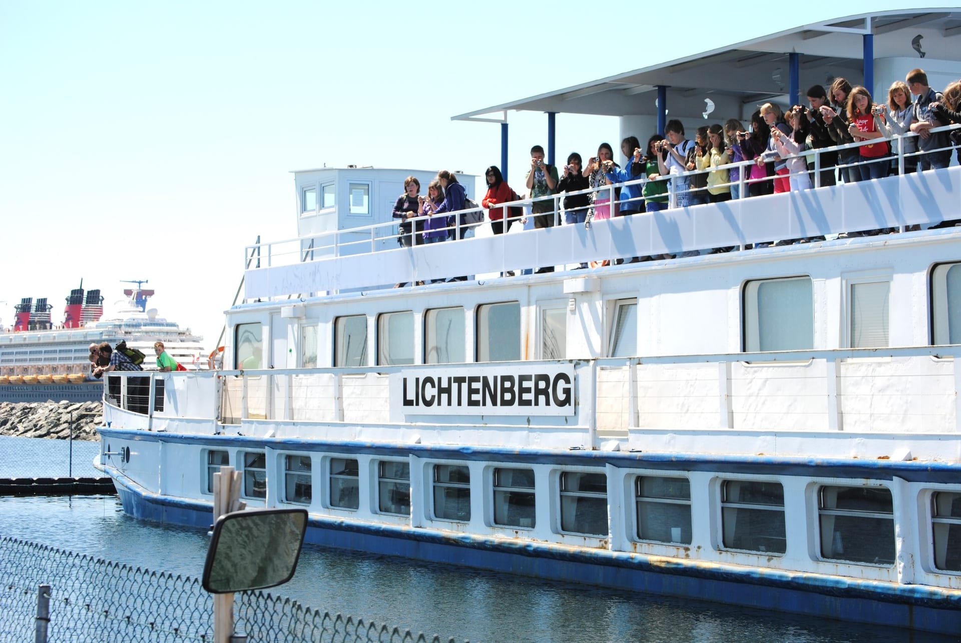 Weißes Fahrgastschiff mit der Aufschrift „LICHTENBERG“. Zahlreiche Passagiere auf dem Oberdeck. Im Hintergrund ein großes Kreuzfahrtschiff und blauer Himmel.