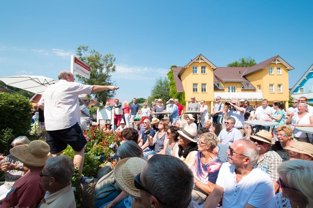 Große Menschenmenge im Freien bei Sonnenschein vor bunten Gebäuden, Menschen lachen und klatschen, fröhliche Stimmung auf einer Veranstaltung.