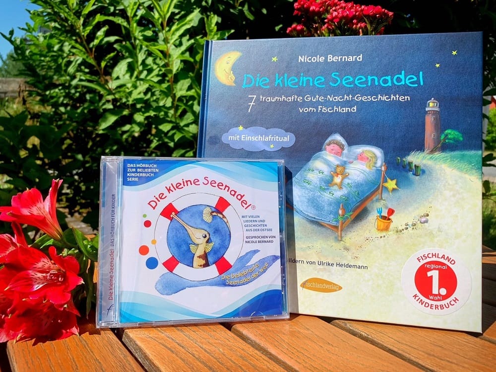 Ein Kinderbuch und ein Hörbuch von Nicole Bernard auf einem Holztisch neben einer roten Blume.