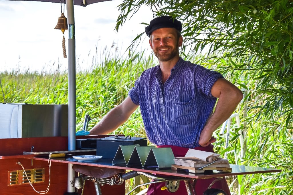 Mann mit blau gestreiftem Hemd und schwarzer Mütze, stehend hinter einem kleinen Outdoor-Stand mit Küchenutensilien, umgeben von hohem Grün.