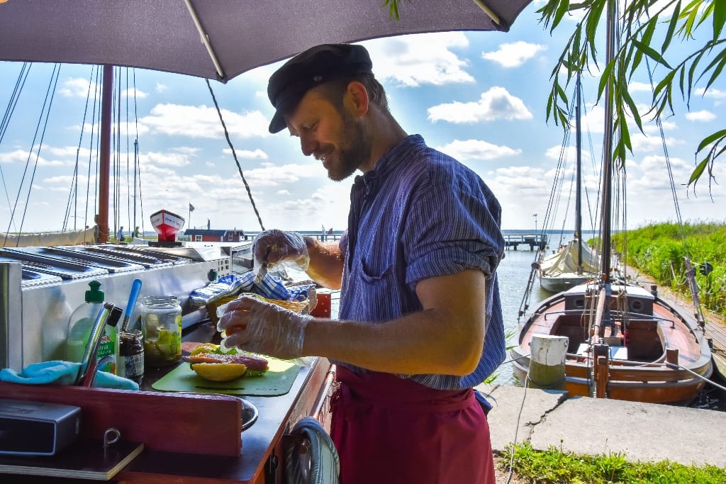 Mann mit blau gestreiftem Hemd und schwarzer Mütze, stehend an einem Outdoor-Stand am Wasser, zubereitet Fischbrötchen, umgeben von Booten und Grün.