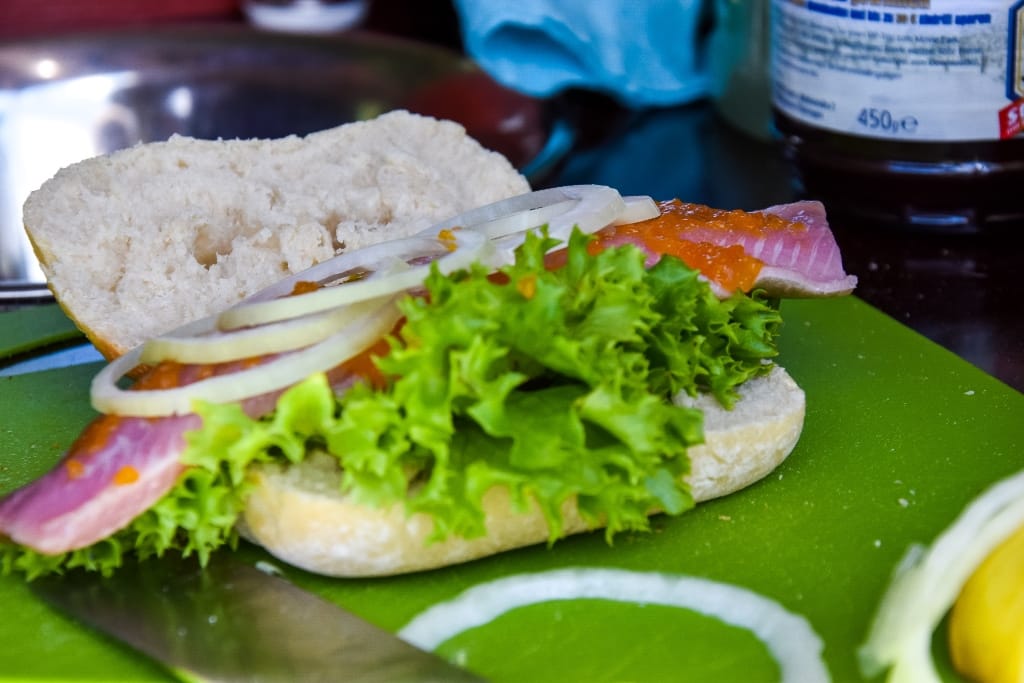 Brötchen mit grünem Salat, Zwiebeln, rohem Fisch und Aufstrich, liegend auf grünem Schneidebrett, im Hintergrund Küchenutensilien.