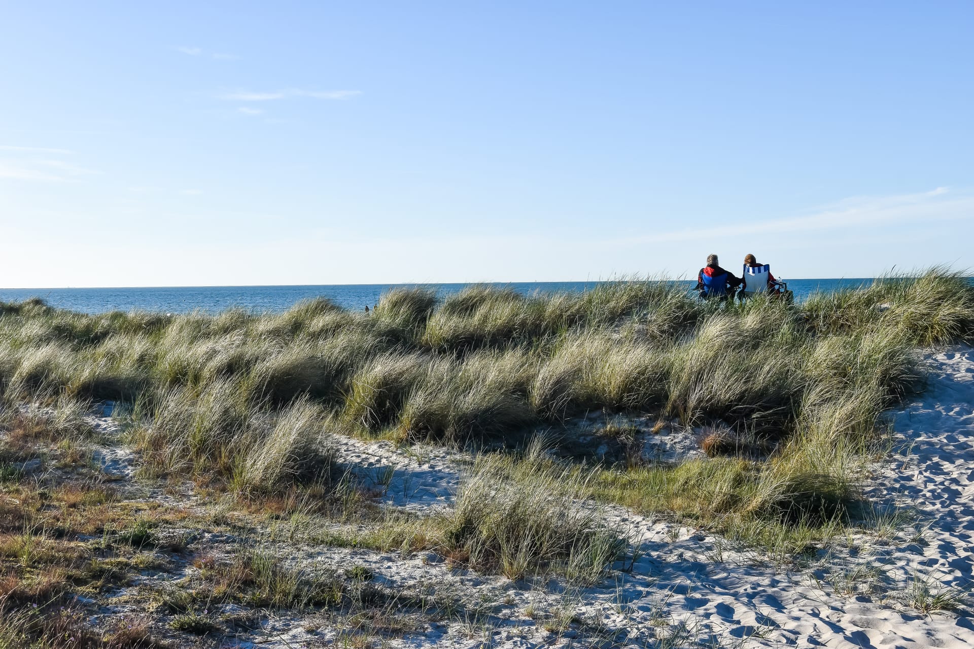 Zwei Personen hinter einer Düne mit Blick auf das Meer unter blauem Himmel.