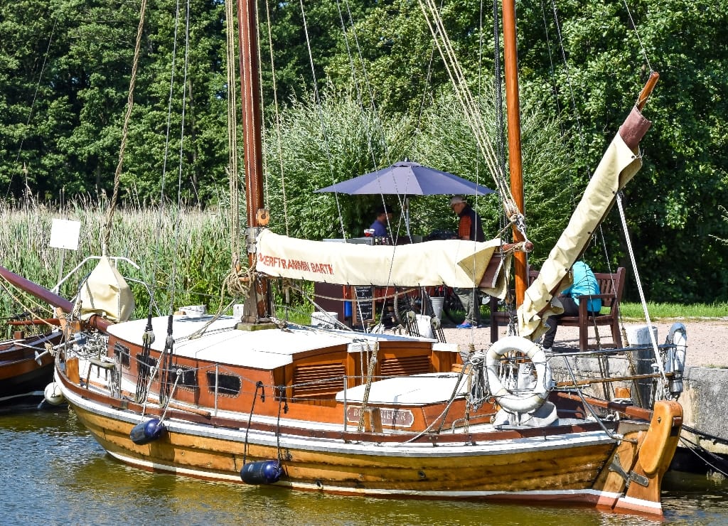 Im Hafen ein vertäutes Holzsegelboot auf dem Wasser, umgeben von grünem Schilf, Bäumen und weiteren Booten.