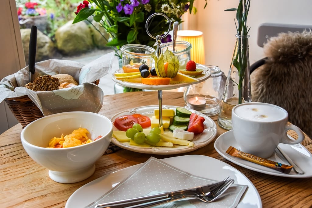 Ein Tisch zum Frühstück gedeckt: Kaffee, ein Brotkorb, eine Etagere mit Käse und Früchten, daneben eine Schüssel mit Cornflakes.