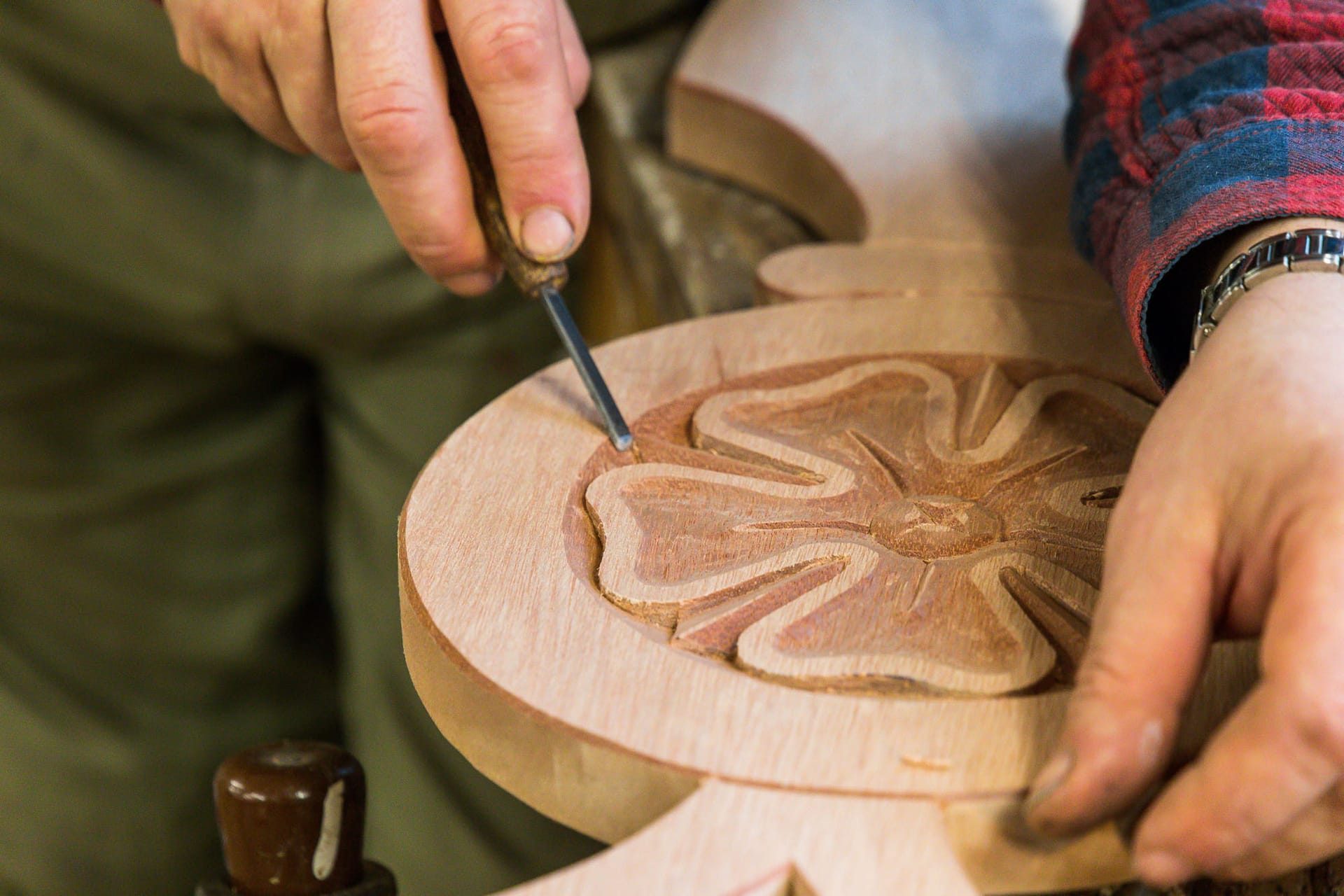 Eine Hand schnitzt mit einem Meißel in Holz; zu sehen ist eine geschnitzte Dekoration in Form einer Blume.