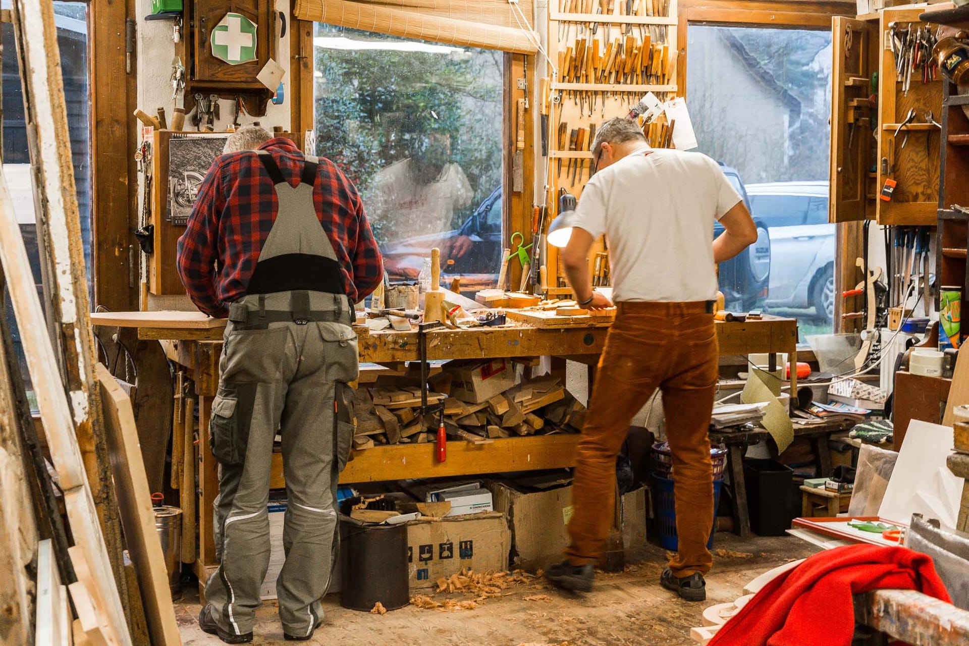 Zwei Männer in einer Holzwerkstatt. Sie arbeiten an Werkbänken. Werkzeuge und Holzmaterialien sind überall sichtbar.
