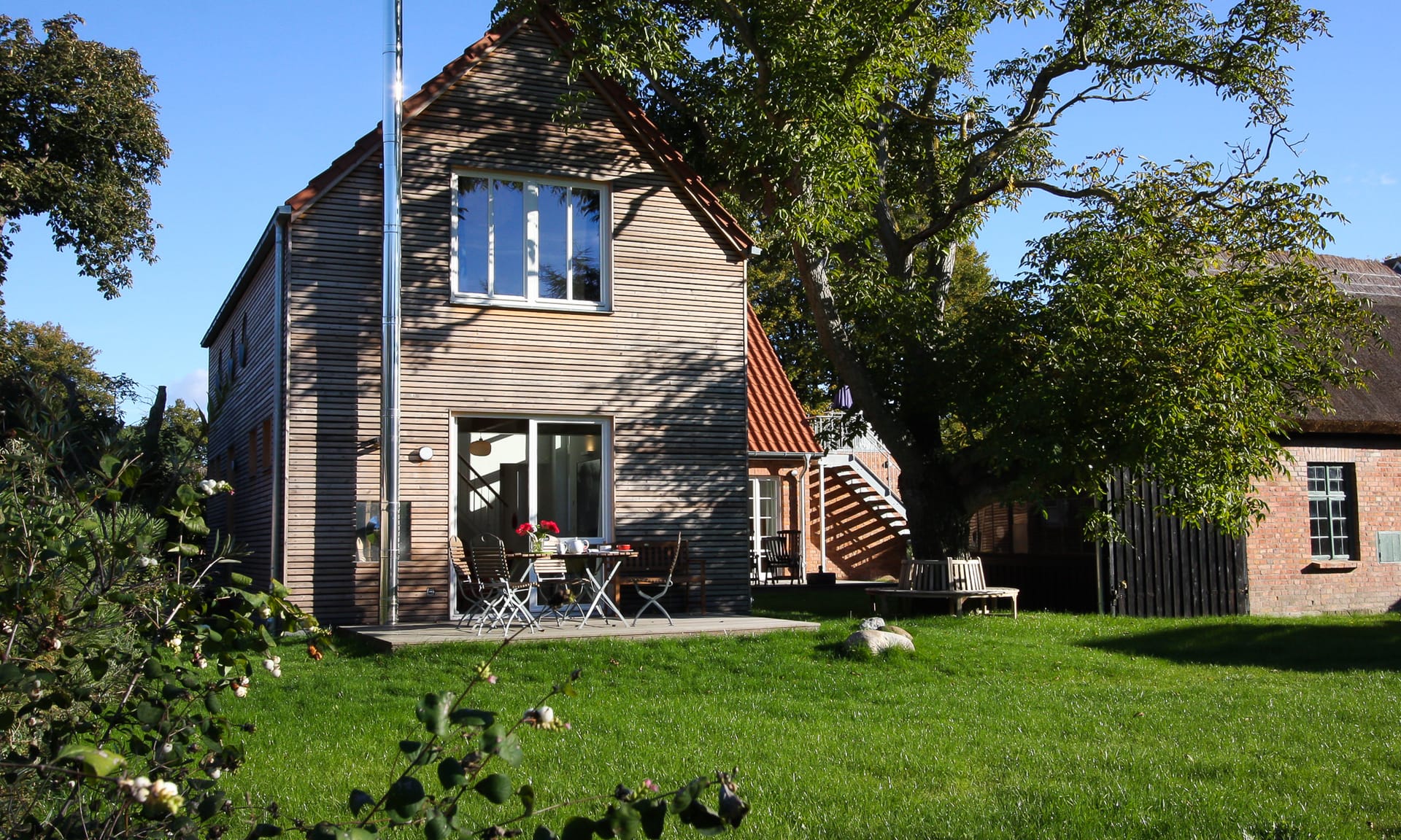 Ein modernes Holzhaus mit Terrasse steht auf einer grünen Wiese neben einem Baum.