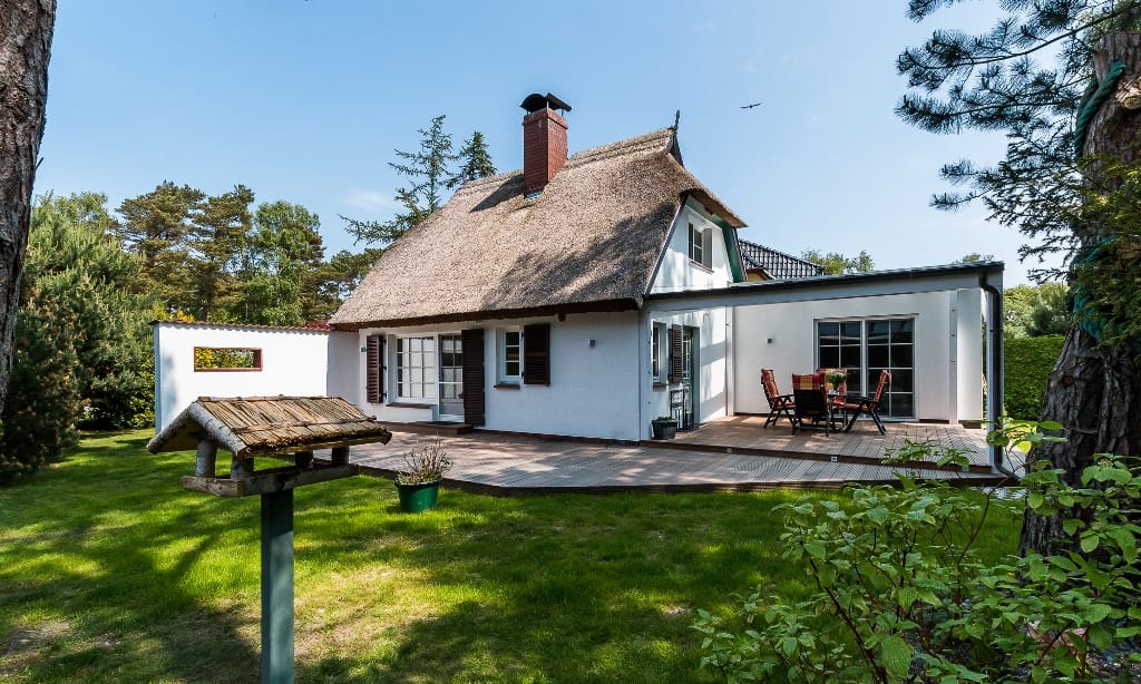 Ein weißes Ferienhaus mit Reetdach und moderner Terrasse steht auf einer grünen Wiese.