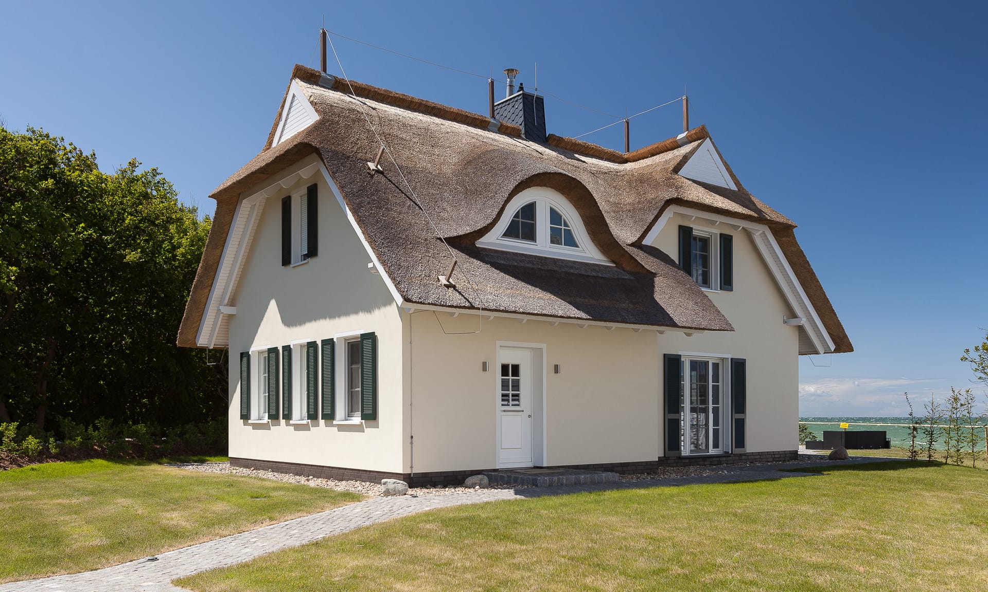 Ein reetgedecktes Ferienhaus mit weißer Fassade, grünen Fensterläden und einem gepflasterten Weg auf einer Rasenfläche unter blauem Himmel.