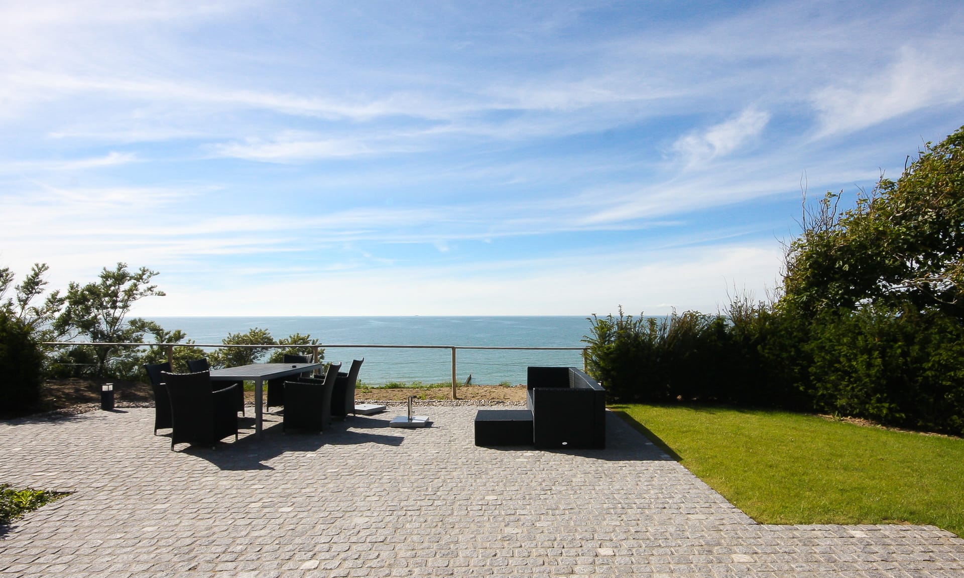 Eine gepflasterte Terrasse mit modernen Gartenmöbeln und freiem Blick auf das Meer unter blauem Himmel.