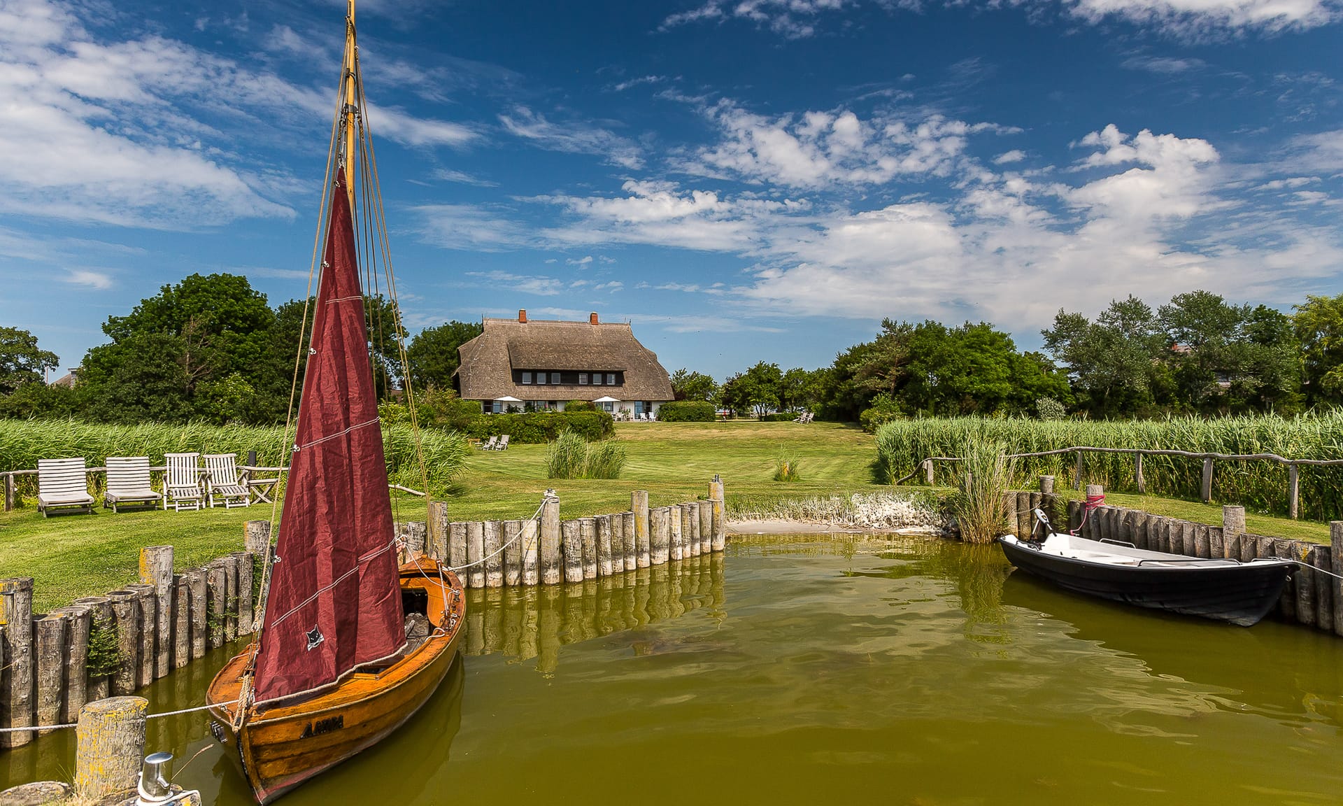Ein historisches Zeesboot und ein Motorboot liegen am hölzernen Steg vor einem weitläufigen Grundstück mit reetgedecktem Haus.