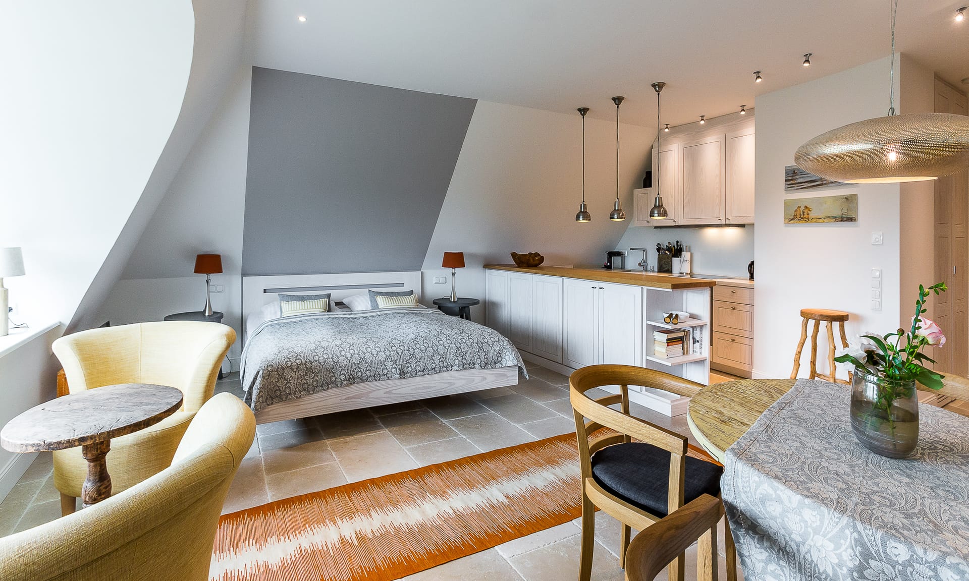 Einzimmerwohnung mit Doppelbett unter einer Dachschräge, integrierter Küchenzeile und offenem Essbereich.