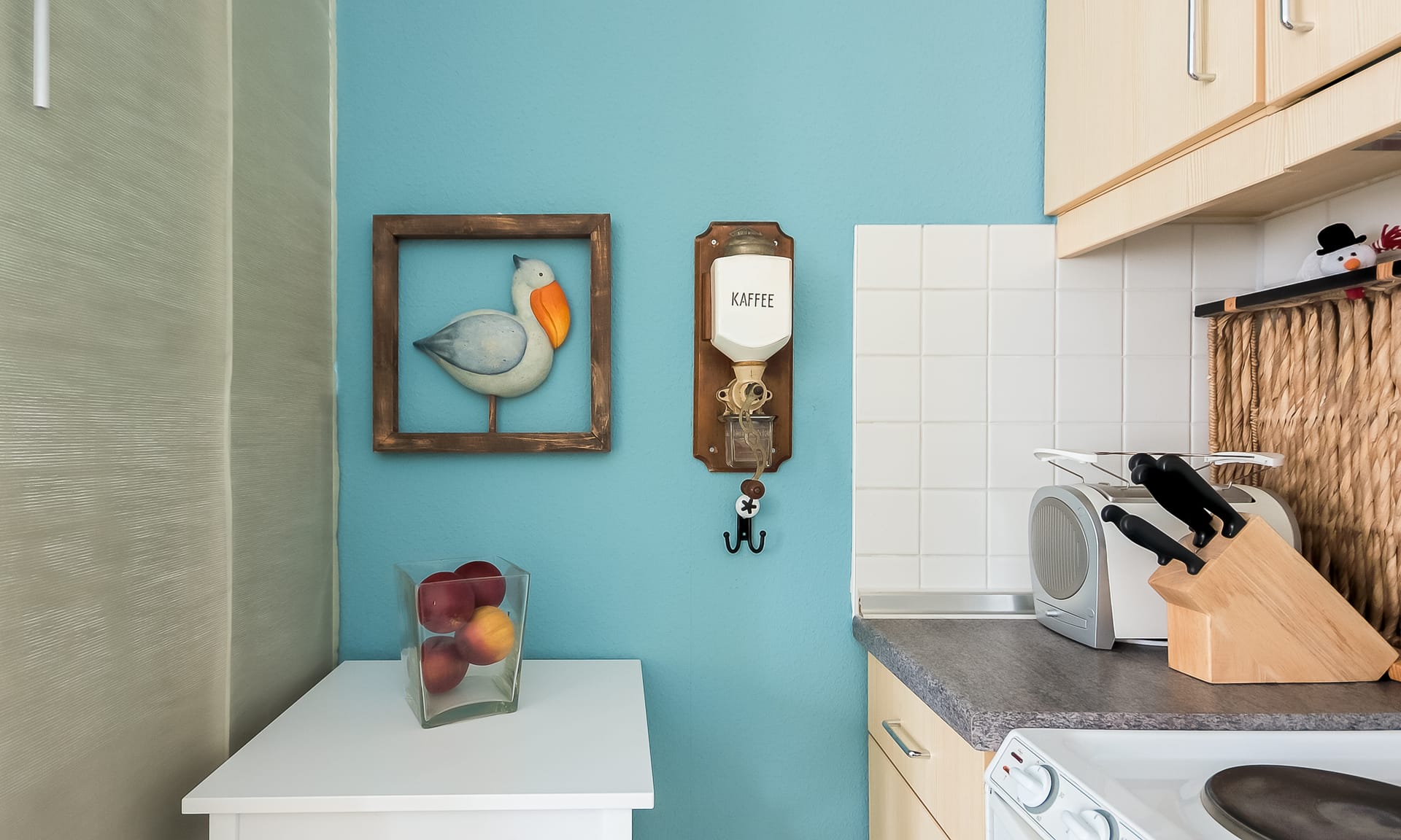 Ein kleiner Küchenbereich mit blauer Wand, einem Messerblock, Toaster, Wandschmuck in Form eines Vogels und einer Kaffeemühle.
