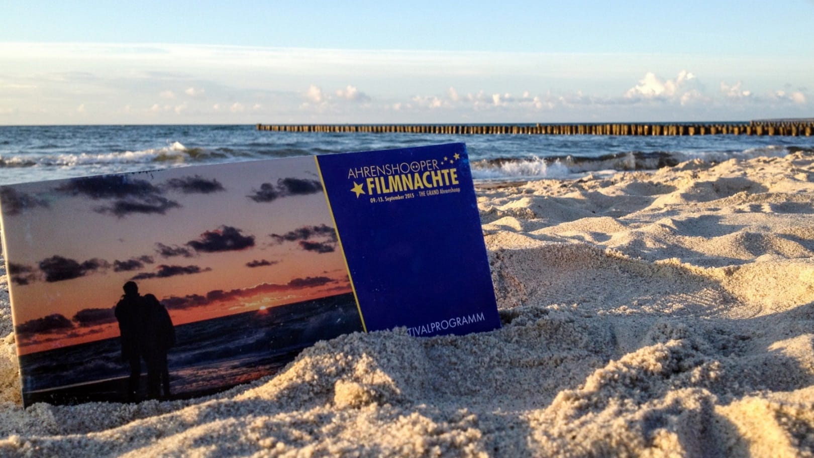 Ein Programmheft der „Ahrenshooper Filmnächte“ im Sand an einem Meeresstrand mit hölzernen Buhnen unter blauem Himmel.