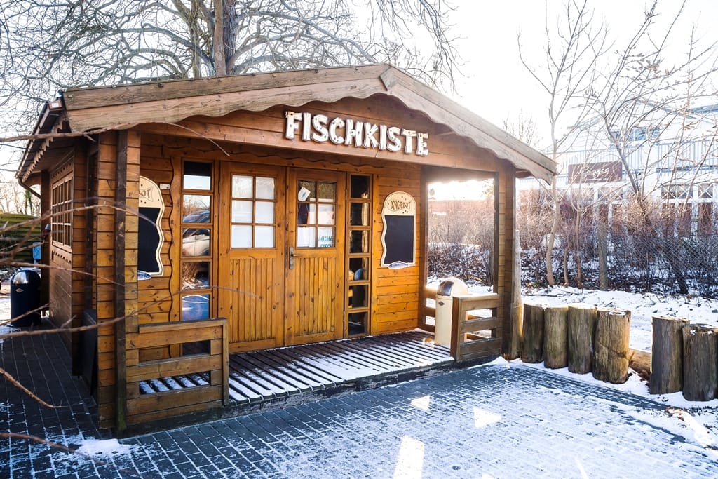 Ein kleines Holzhaus mit der Aufschrift „FISCHKISTE“ in einer verschneiten Außenanlage.