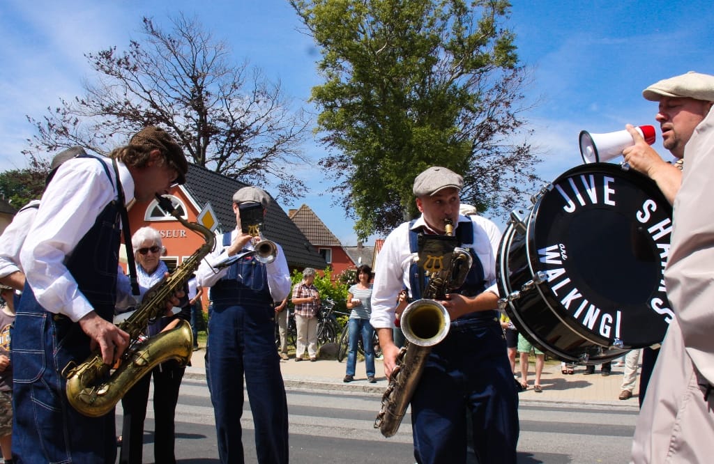 Eine Blaskapelle aus mehreren Musikern spielt mit Saxophonen, einer Trompete und einer Basstrommel auf einer Straße vor Zuschauern im Freien.