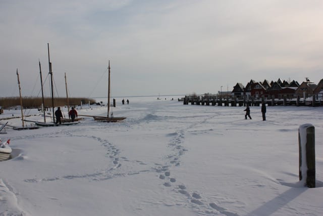 Gefrorener Hafen mit viel Schnee. Menschen gehen auf der gefrorenen Wasseroberfläche. Im Hintergrund Steg, verschneite Gebäude und Boote.