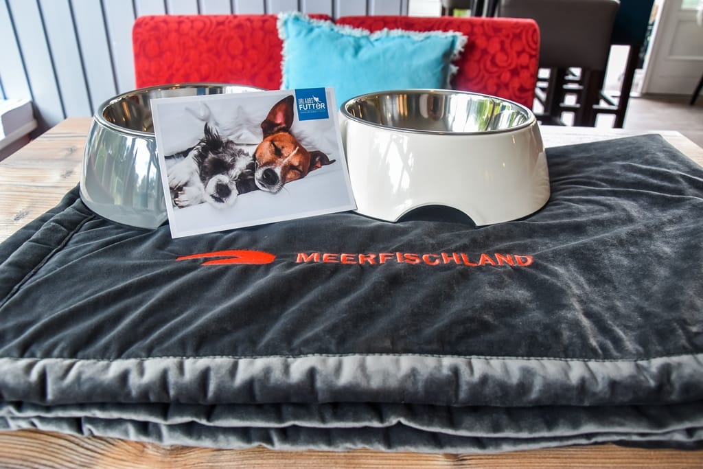 Eine dunkelgraue Hundedecke mit der Aufschrift "Meerfischland" liegt neben zwei Futternäpfen und einer Grußkarte auf einem Holztisch.