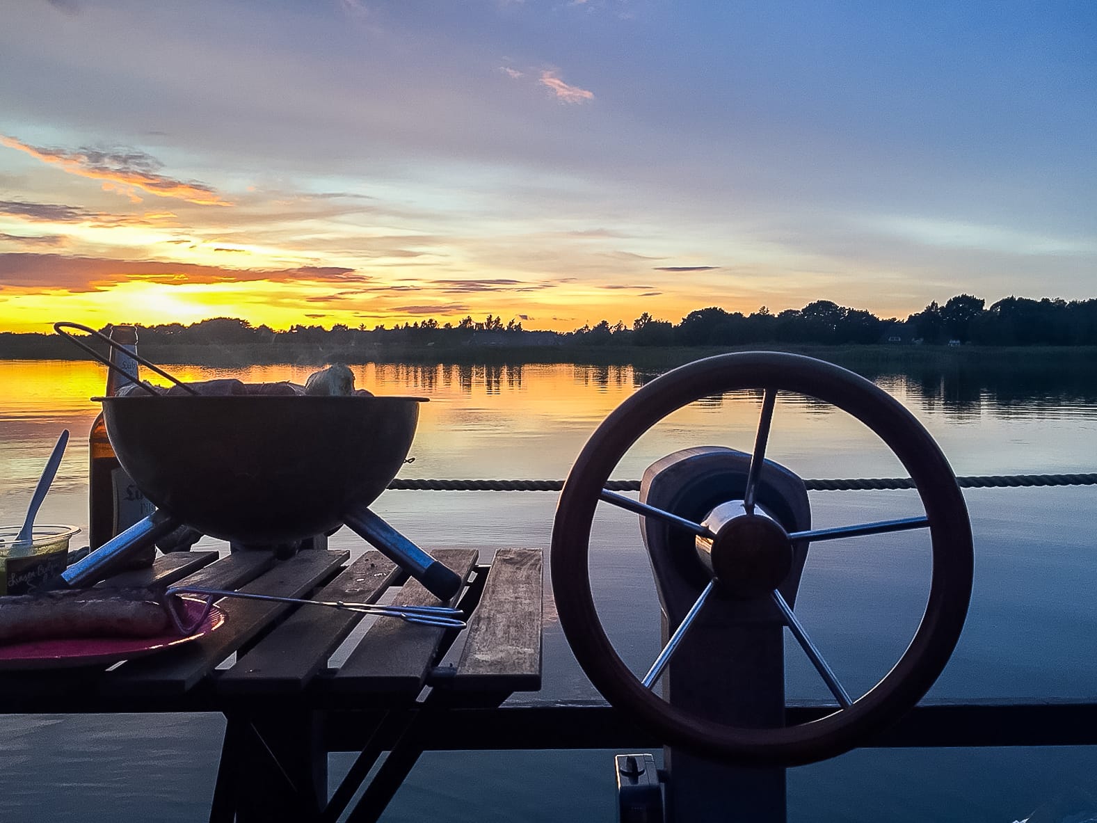 Blick auf das Floßruder, daneben ein Grill, im Hintergrund Sonnenuntergang.