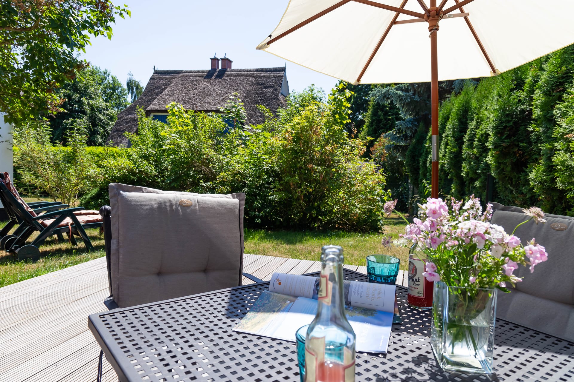 Ein gedeckter Tisch mit Blumen steht auf einer sonnigen Holzterrasse vor einem Reetdachhaus.