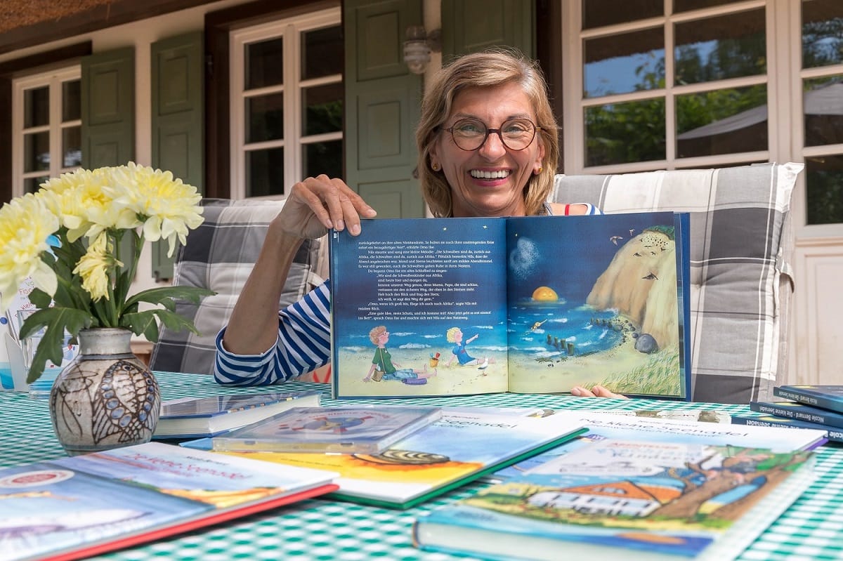 Eine lächelnde Frau mit Brille am Tisch mit Kinderbüchern und einer Vase mit gelben Blumen.