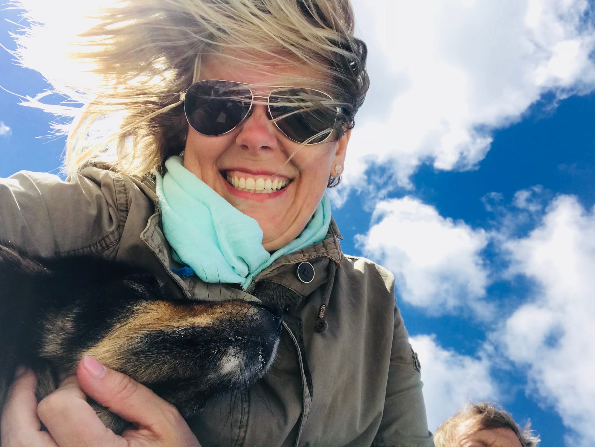 Frau hält einen Hund im Arm, im Hintergrund blauer Himmel mit Wolken.