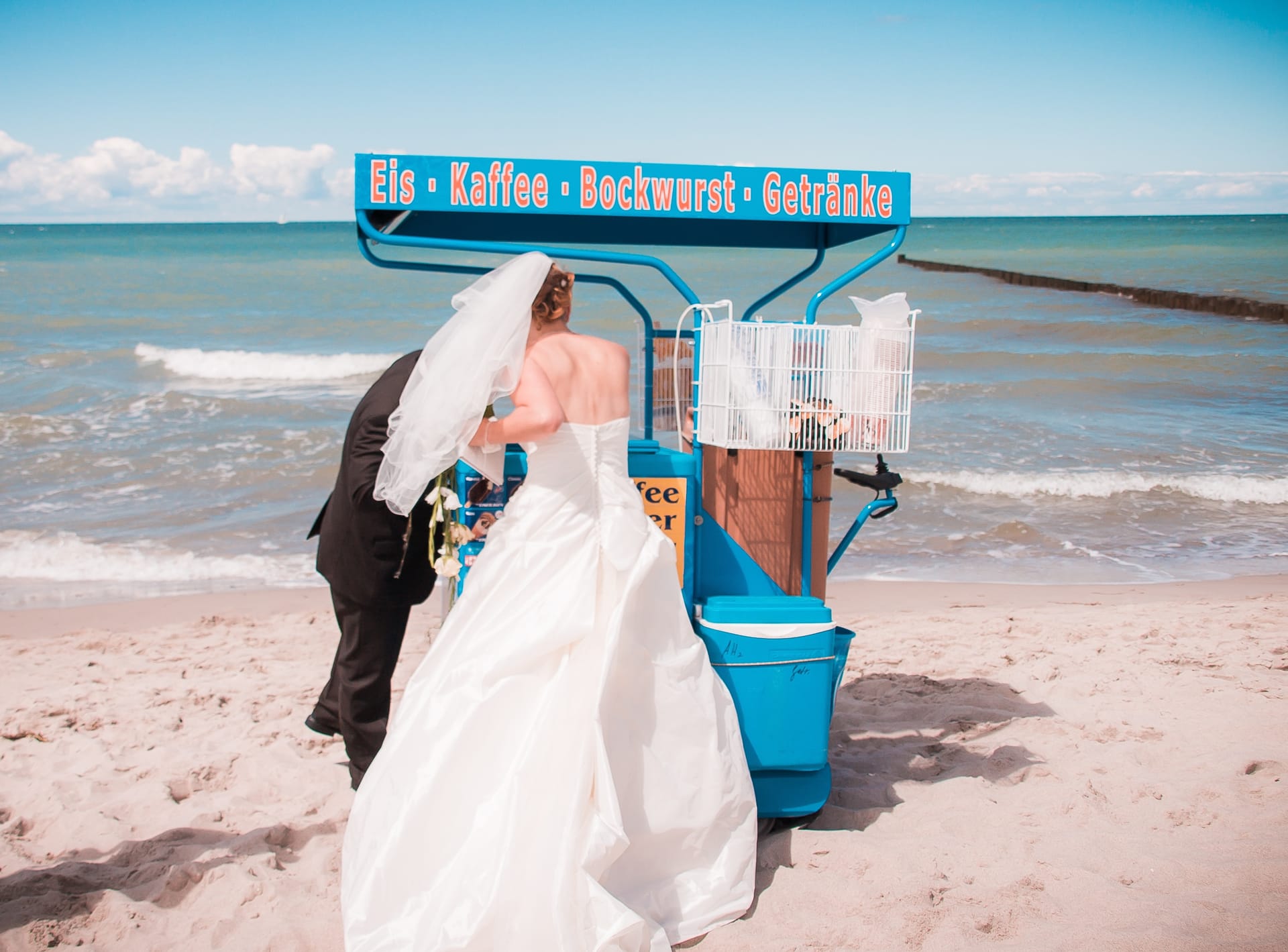 Ein Brautpaar in Hochzeitskleidung an einem Strand und kauft etwas an einem kleinen Imbisswagen. Im Hintergrund sieht man das Meer und den Sandstrand.