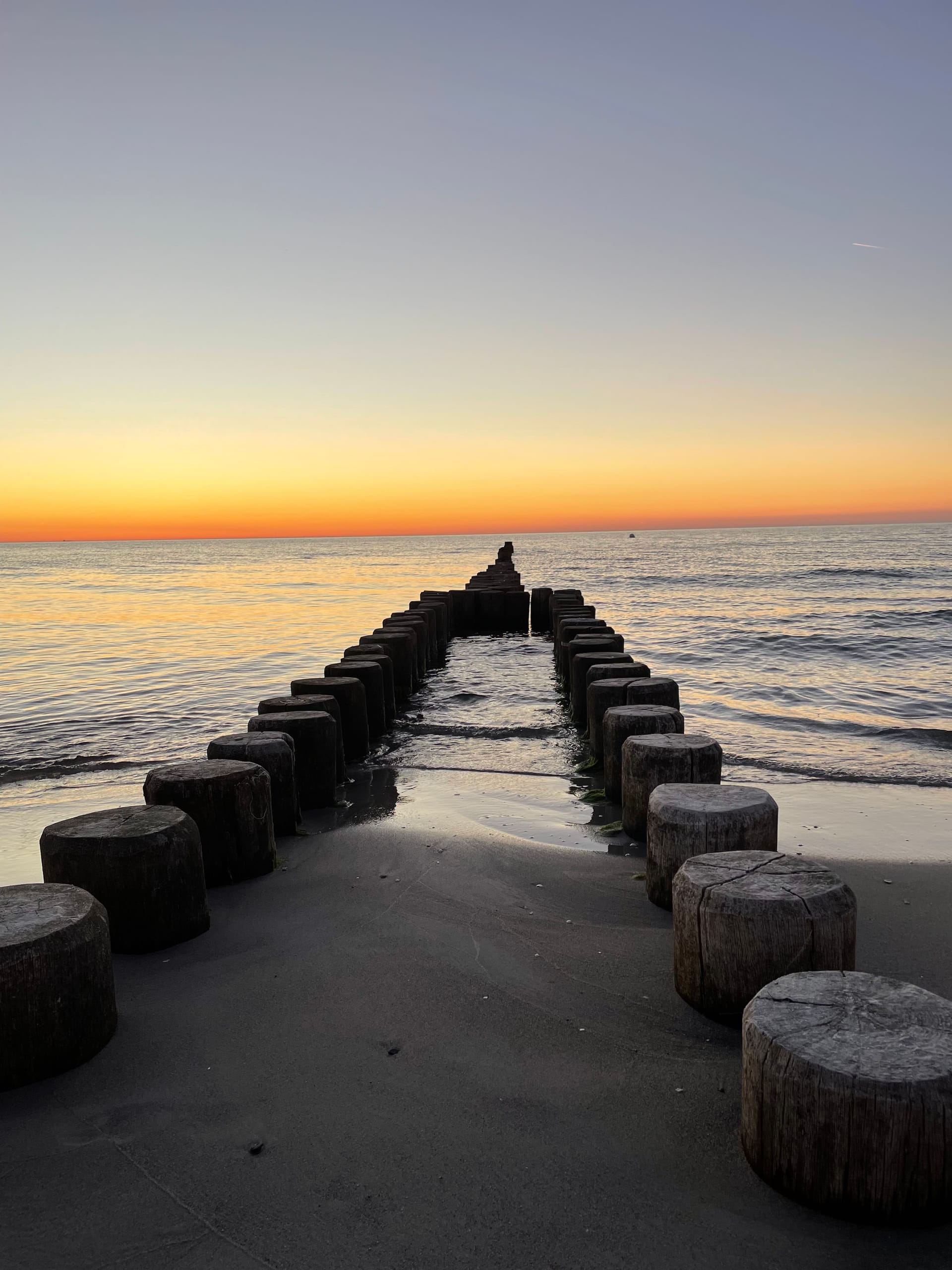 Strand mit hölzernen Pfählen, die vom Ufer ins Meer führen, bei Sonnenuntergang.