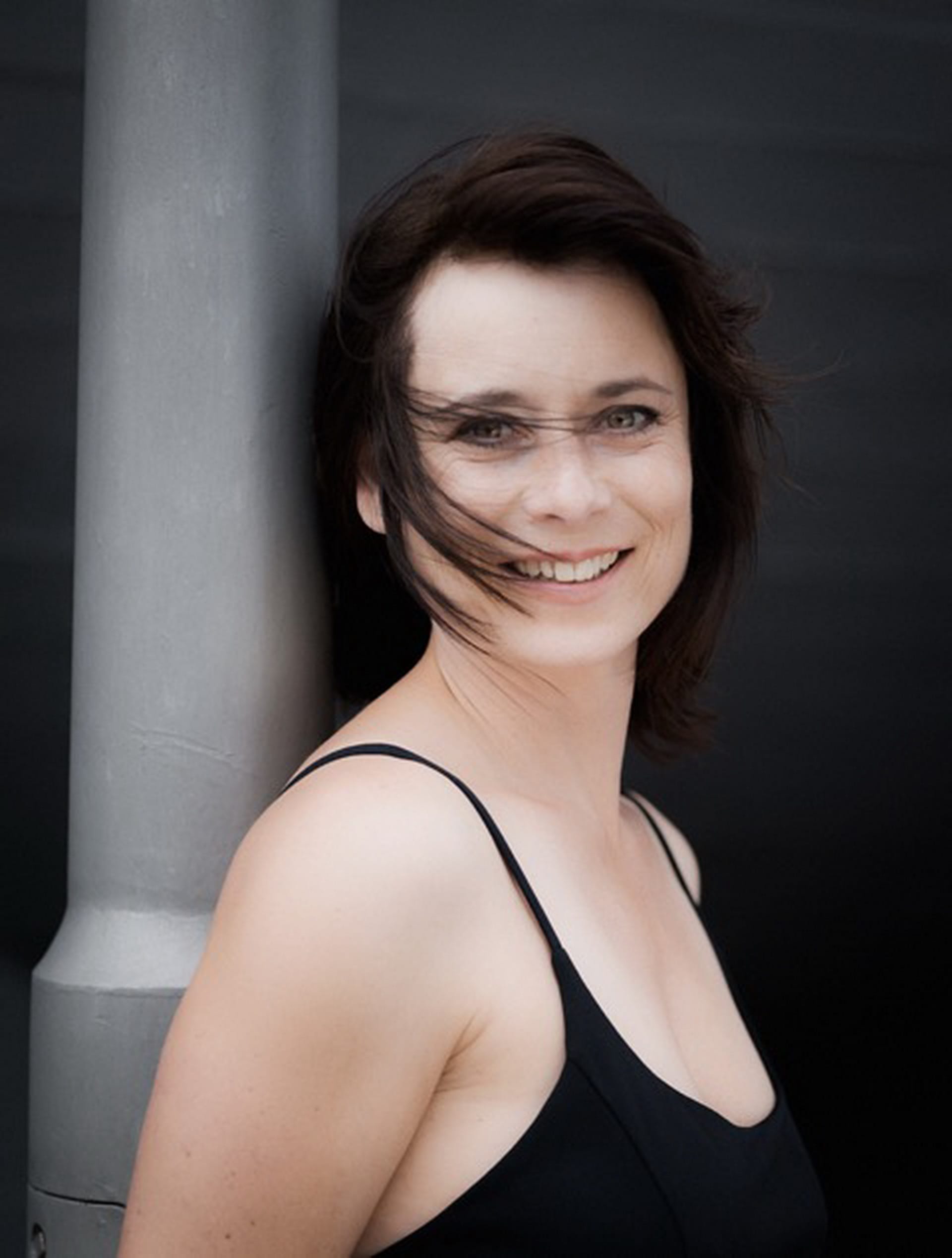 Frau mit schwarzen Haaren in schwarzem Kleid, lachend und in die Kamera blickend.