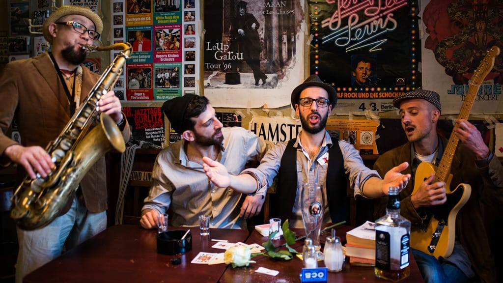 Vier Musiker mit ihren Instrumenten posieren an einem Tisch in einer Bar vor einer Wand voller Plakate.