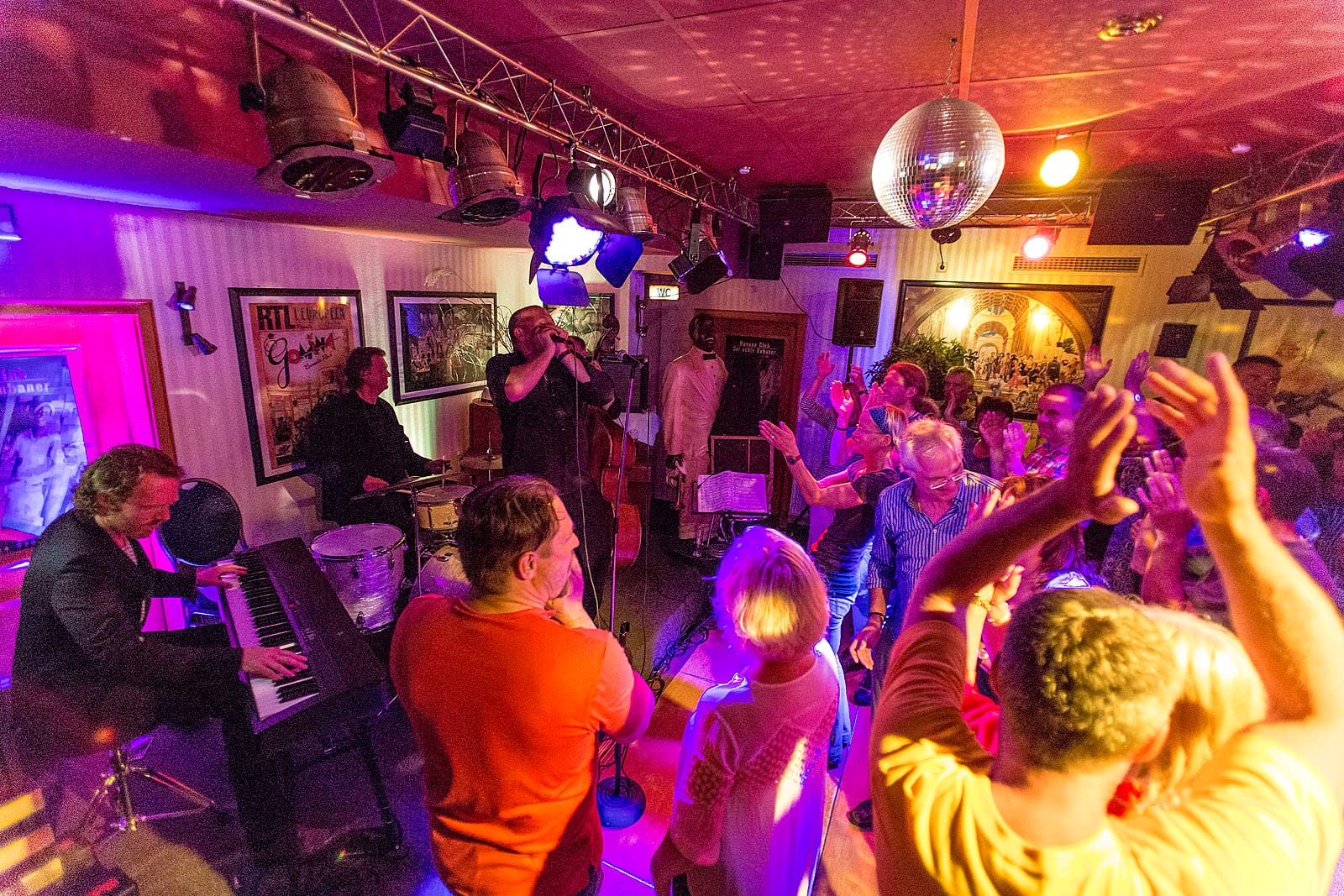 Eine Band spielt live Musik auf einer Bühne vor einer tanzenden Menschenmenge in einem beleuchteten Raum.