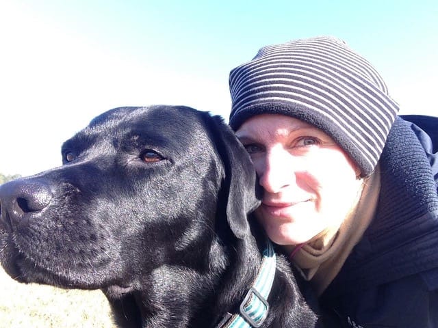 Eine lächelnde Frau mit gestreifter Mütze und ein schwarzer Labrador machen ein gemeinsames Foto im Freien.