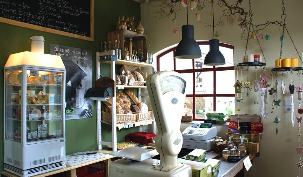 Innenraum eines kleinen Ladens. Auf der Theke eine Retro-Waage und Kasse, dahinter ein Regal mit Brot, Gebäck und Flaschen. Hinten links eine Vitrine mit Lebensmitteln.