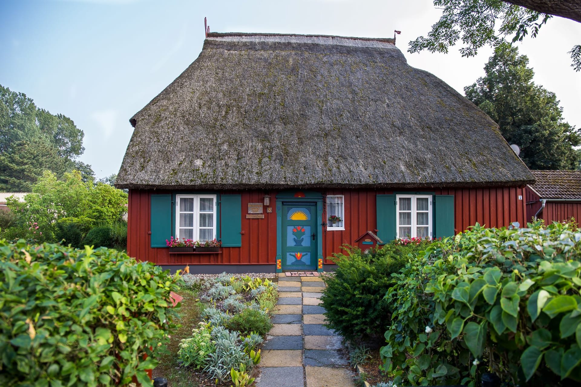Rotes Haus mit Reetdach, umgeben von Gartenpflanzen. Es hat eine blaue Tür und grüne Fensterläden.