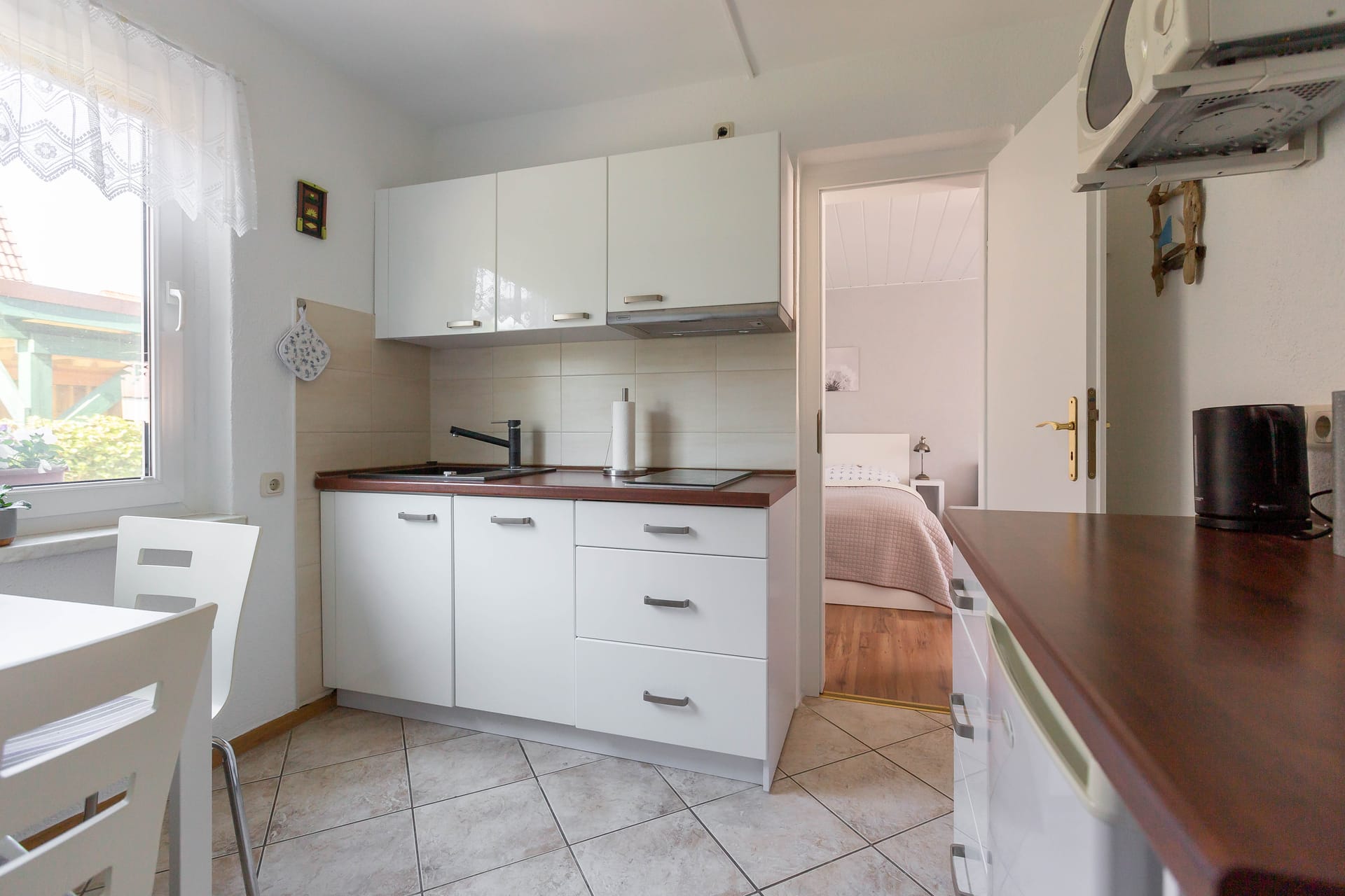 Eine weiße Einbauküche mit dunkler Arbeitsplatte, Ceranfeld und Spüle sowie Blick in das angrenzende Schlafzimmer.