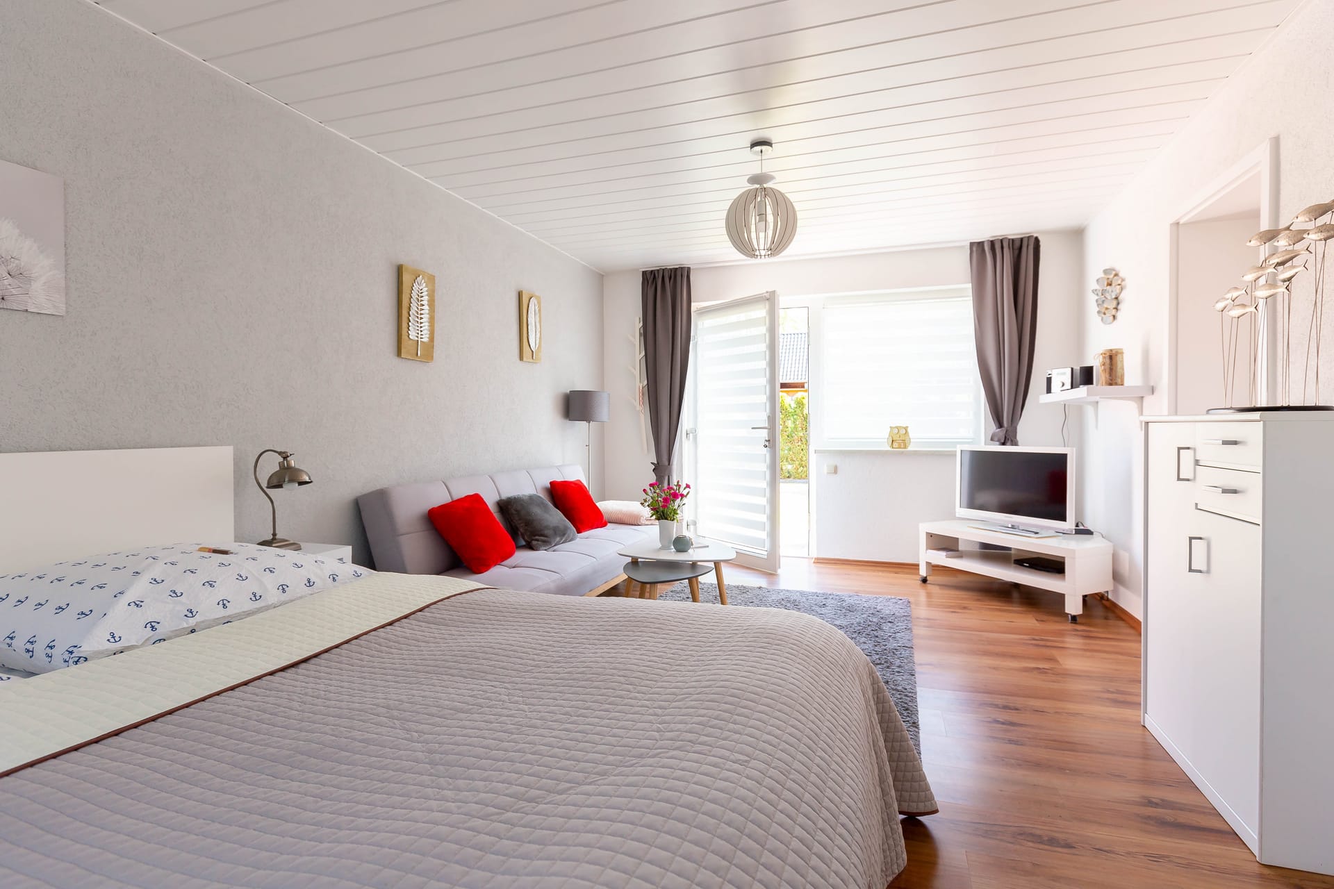 Helles Wohn- und Schlafzimmer mit Doppelbett, grauem Sofa, TV-Schrank und direktem Zugang zum sonnigen Außenbereich.