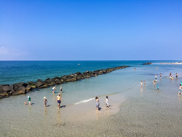 Strand bei sonnigem Wetter, mit Menschen im Meer, Wellenbrecher und blauem Himmel.